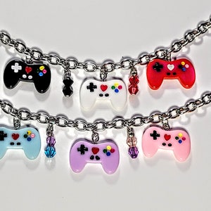 May include: A silver chain bracelet with six colorful game controller charms. Each charm features a different color, including black, white, red, light blue, purple, and pink. The charms have a heart, plus sign, and colored circles on them.