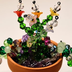 May include: A handcrafted beaded flower arrangement in a small terracotta pot. The flowers are made of clear, red, yellow, pink, and purple beads. The leaves are green, and the pot is filled with purple and clear stones. The arrangement is a unique decorative item.