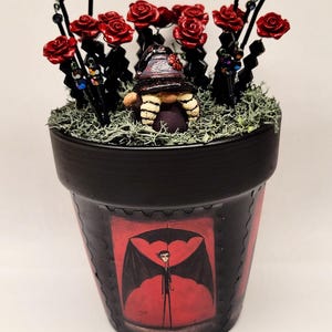 May include: A black flower pot featuring a graphic of a figure with bat wings holding an umbrella against a red background. The pot contains green moss, a small gnome figurine with a dark hat, and several black stems topped with red roses.