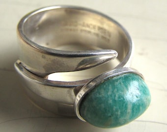 May include: A silver ring with a large, oval, turquoise-colored stone. The ring has a unique, open design with a split band that wraps around the finger. The stone is set in a silver bezel.