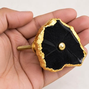 May include: A cabinet knob featuring a flat, irregular black stone face with a textured, metallic gold-colored edge. A small, smooth gold-colored metal sphere is centered on the black surface, and a threaded gold-colored metal bolt extends from the back.