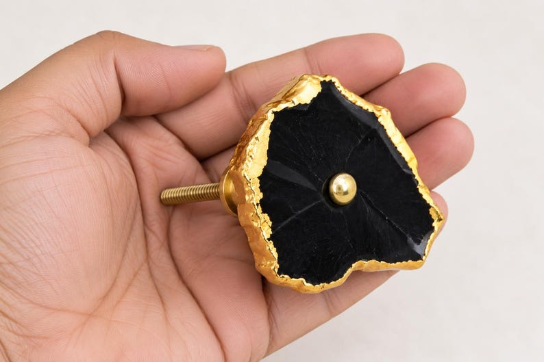 May include: A cabinet knob featuring a flat, irregular black stone face with a textured, metallic gold-colored edge. A small, smooth gold-colored metal sphere is centered on the black surface, and a threaded gold-colored metal bolt extends from the back.