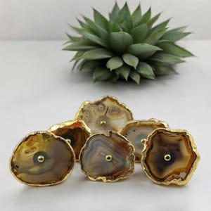 Pode incluir: Cinco puxadores de gaveta de &aacute;gata com acabamento dourado. A &aacute;gata &eacute; uma pedra natural com redemoinhos marrons, brancos e cinzentos. Os puxadores s&atilde;o uma adi&ccedil;&atilde;o &uacute;nica e elegante a qualquer casa.