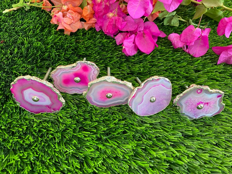 May include: Five pink agate drawer pulls with silver metal accents. The pulls are arranged on a green artificial grass surface.