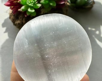 May include: A white selenite charging plate with a smooth, polished surface and subtle striations. The plate is round and has a slightly domed shape.