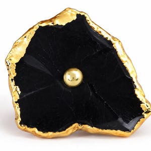 May include: A decorative cabinet knob featuring an irregular, flat black stone slice with a polished, reflective surface. The edges of the stone are encased in a thick, textured gold-tone metal trim, and a small, smooth gold-tone metal sphere is centered.