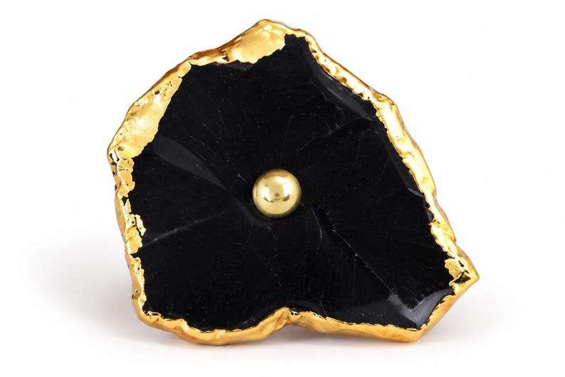 May include: A decorative cabinet knob featuring an irregular, flat black stone slice with a polished, reflective surface. The edges of the stone are encased in a thick, textured gold-tone metal trim, and a small, smooth gold-tone metal sphere is centered.