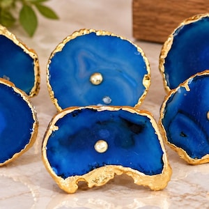 May include: A set of five blue agate stone drawer knobs with irregular, organic shapes and natural banding patterns. Each knob features a polished, vibrant blue surface with a metallic gold-toned electroplated edge and a central gold-toned metal bolt.