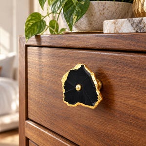 May include: A drawer knob featuring a black, irregularly shaped stone slice with a metallic gold-toned edge and a central gold-toned circular fastener, mounted on a wooden drawer front with a visible horizontal wood grain pattern.