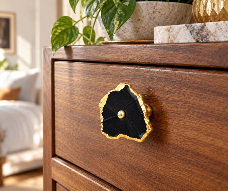 May include: A drawer knob featuring a black, irregularly shaped stone slice with a metallic gold-toned edge and a central gold-toned circular fastener, mounted on a wooden drawer front with a visible horizontal wood grain pattern.