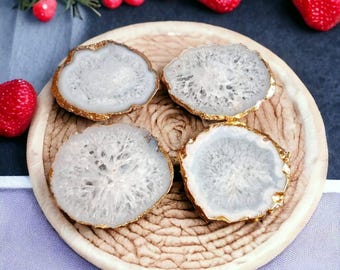 May include: Four circular agate slices with translucent white centers and textured crystalline patterns, each featuring a metallic gold-toned electroplated edge. These stone coasters are arranged on a round, woven, light tan textured base.
