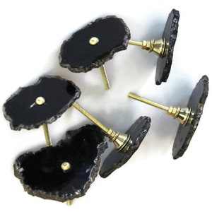 May include: A set of four cabinet knobs featuring irregular, flat, black stone slices with natural, rough, metallic-toned edges. Each knob has a central gold-toned metal screw post and a matching gold-toned circular base plate for mounting.