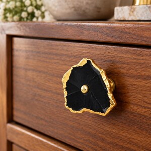 May include: A drawer knob featuring a flat, irregular black stone slice with a textured, metallic gold-toned edge. The center of the black stone is accented with a small, smooth, round gold-toned metal stud that secures the piece to the wooden drawer.