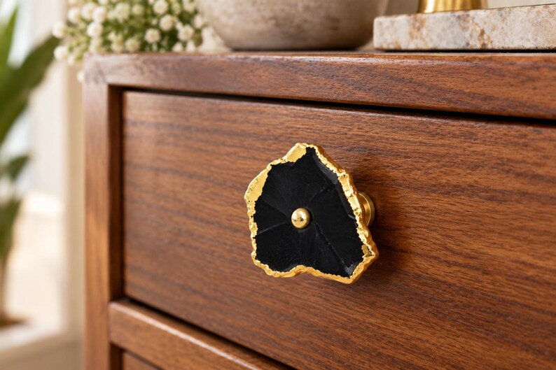 May include: A drawer knob featuring a flat, irregular black stone slice with a textured, metallic gold-toned edge. The center of the black stone is accented with a small, smooth, round gold-toned metal stud that secures the piece to the wooden drawer.