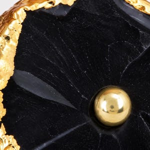 May include: A cabinet knob featuring a polished black stone slice with a natural, irregular shape. The edges of the stone are finished with a textured, metallic gold-tone coating. A small, smooth, gold-tone metal sphere is centered on the black stone.