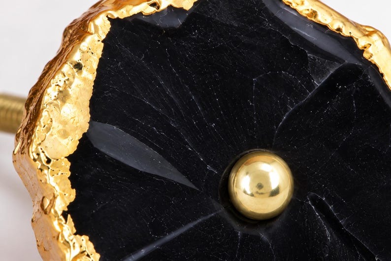 May include: A cabinet knob featuring a polished black stone slice with a natural, irregular shape. The edges of the stone are finished with a textured, metallic gold-tone coating. A small, smooth, gold-tone metal sphere is centered on the black stone.