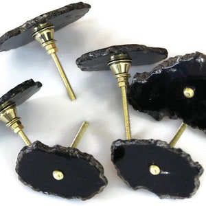 May include: A set of five drawer knobs featuring irregular, polished black agate slices with natural, rough-textured edges. Each knob is attached to a gold-toned metal bolt and hardware, providing a functional handle for furniture or cabinetry.