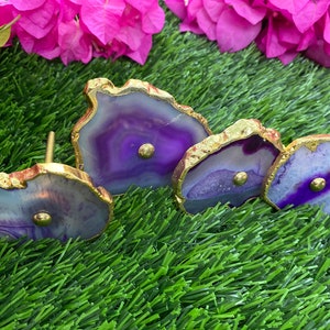 May include: Three purple agate knobs with gold trim. The knobs are different sizes and have a natural, irregular shape. They are set against a green grass background.