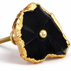 May include: A cabinet knob featuring an irregular, flat black stone face with a textured, metallic gold-colored edge. A small, smooth, gold-colored metal sphere is centered on the black surface. A threaded gold-colored metal bolt extends from the back.