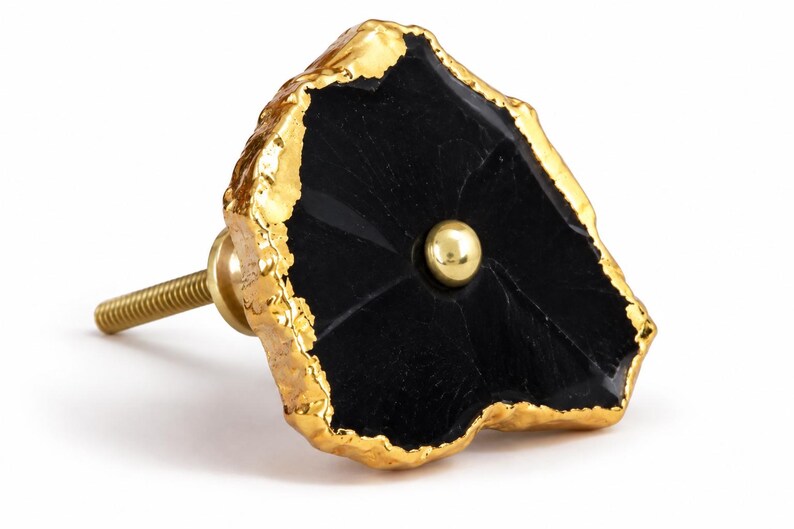 May include: A cabinet knob featuring an irregular, flat black stone face with a textured, metallic gold-colored edge. A small, smooth, gold-colored metal sphere is centered on the black surface. A threaded gold-colored metal bolt extends from the back.