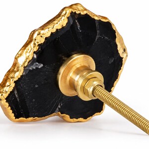 May include: A cabinet knob featuring a slice of black stone with a textured, metallic gold-colored edge. The back of the knob includes a brass-toned metal mounting base with a threaded bolt extending outward for installation on furniture or doors.