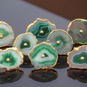 May include: A set of seven green agate drawer pulls with gold trim. Each pull features a unique pattern of green and white agate with a gold dot in the center.
