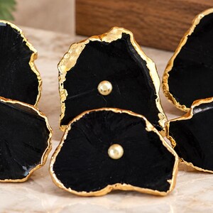 May include: A set of five black stone drawer knobs with irregular, organic shapes. Each knob features a polished black surface with a metallic gold-toned edge and a small, centered gold-toned spherical accent piece for mounting purposes.