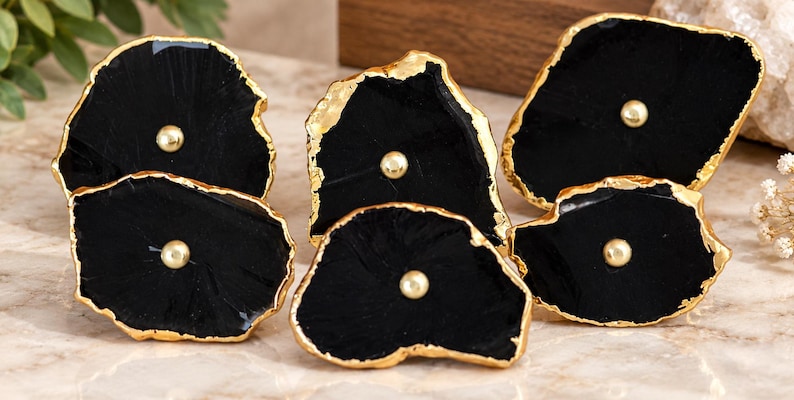 May include: A set of five black stone drawer knobs with irregular, organic shapes. Each knob features a polished black surface with a metallic gold-toned edge and a small, centered gold-toned spherical accent piece for mounting purposes.