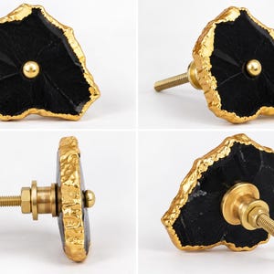 May include: A cabinet knob featuring a flat, irregular black stone face with a textured, metallic gold-colored edge. A small gold-colored metal sphere is centered on the front, and a threaded gold-colored metal bolt extends from the back.