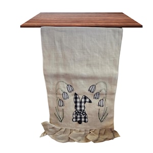 Op de afbeelding: Een beige linnen tafelloper met een geborduurd paashaasontwerp. De haas is zwart-wit geruit, omringd door geborduurde bloemen. De onderkant heeft een gefranjerde rand.