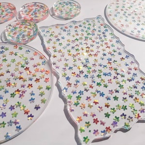May include: A set of six clear resin coasters with a scattered rainbow glitter design. The coasters are round and have a scalloped edge.
