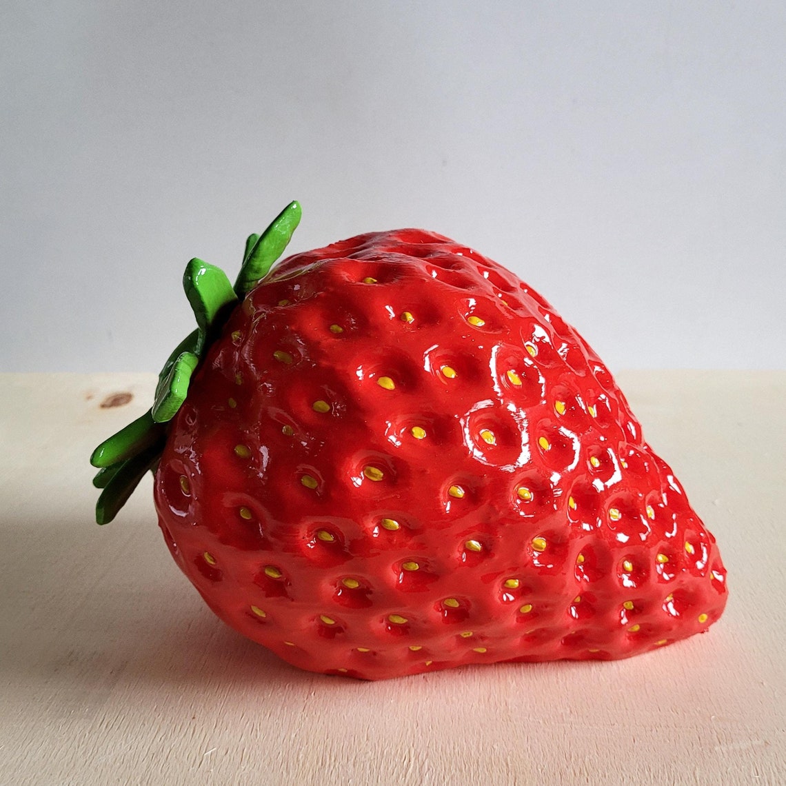 May include: A red and green ceramic strawberry with yellow dots. The strawberry is a realistic depiction of a strawberry and has a green stem and leaves.