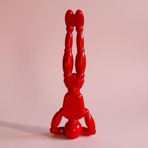 Könnte beinhalten: Eine glänzend rote Skulptur einer Figur im Handstand. Die Figur hat abgerundete Gliedmaßen und einen kugelförmigen Kopf und eine Basis. Der Hintergrund ist ein zartes Rosa.