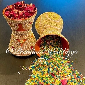 May include: Two decorative orange bowls with gold and white embellishments. One bowl is filled with colorful rice and rose petals. The bowls are on a dark brown surface.