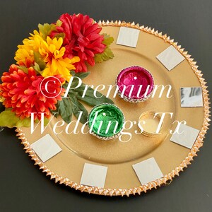 May include: A gold tray with a decorative border of small gold triangles and a floral arrangement of red, yellow, and green flowers. The tray has six small silver squares and three small bowls, one pink, one green, and one gold.