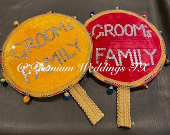 May include: Two round, decorative hand-held fans with gold trim. One fan is yellow with the text "Groom's Family" in silver sequins. The other fan is red with the text "Groom's Family" in silver sequins. Both fans have gold tassels.