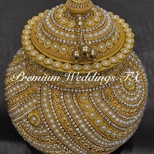 May include: A gold-colored, round decorative box with a lid, heavily embellished with pearls and rhinestones. The box features intricate patterns and a tassel detail. The text "Premium Weddings FX" is visible on the front of the box.