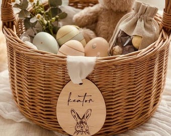 May include: A round woven wicker basket with a handle, containing a plush tan rabbit, several pastel-colored eggs, and a small drawstring pouch. An oval wooden tag hangs from the front, featuring the name Keaton and a line-art rabbit illustration.