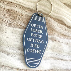 May include: A blue motel-style keychain with a silver metal ring. The keychain features white engraved text that reads: GET IN LOSER, WE'RE GETTING ICED COFFEE. The design includes white horizontal line accents at the top and bottom of the tag.