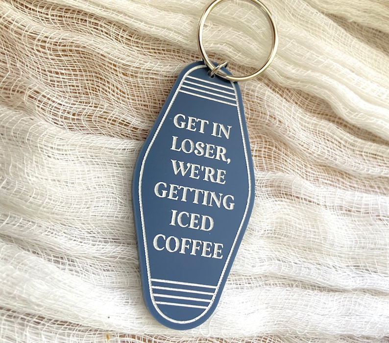 May include: A blue motel-style keychain with a silver metal ring. The keychain features white engraved text that reads: GET IN LOSER, WE'RE GETTING ICED COFFEE. The design includes white horizontal line accents at the top and bottom of the tag.