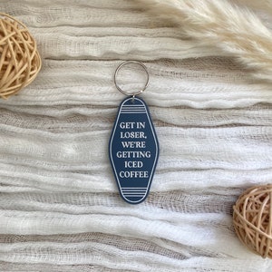 May include: A dark blue motel-style keychain with a silver metal ring. The keychain has a pointed oval shape with white border lines and the white text: GET IN LOSER, WE'RE GETTING ICED COFFEE. The design is centered and features a clean, bold font.