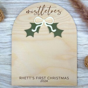 Puede incluir: Un letrero de madera con la parte superior redondeada y un dise&ntilde;o de mu&eacute;rdago verde. El letrero dice "Mistletoes" y "Rhett's First Christmas 2024".