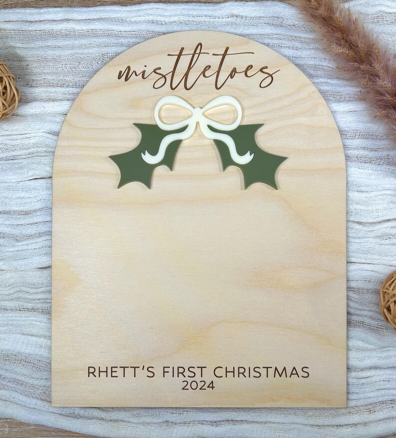 Puede incluir: Un letrero de madera con la parte superior redondeada y un dise&ntilde;o de mu&eacute;rdago verde. El letrero dice "Mistletoes" y "Rhett's First Christmas 2024".