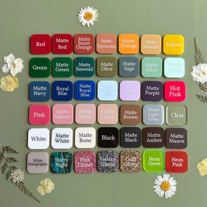 May include: A collection of square acrylic color swatches arranged in a grid, each labeled with its respective color name including matte, glitter, and neon finishes. The swatches are displayed on a solid green background with white flower accents.