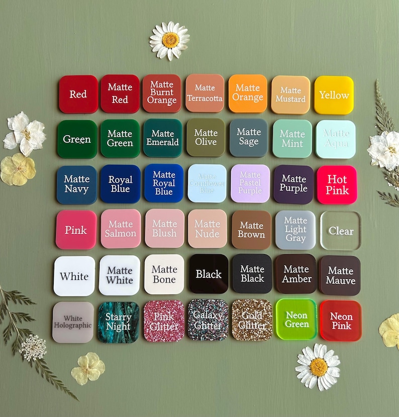 May include: A collection of square acrylic color swatches arranged in a grid, each labeled with its respective color name including matte, glitter, and neon finishes. The swatches are displayed on a solid green background with white flower accents.