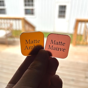 May include: Two square acrylic swatches with rounded corners. The left swatch is translucent amber with the text Matte Amber printed in black. The right swatch is translucent mauve with the text Matte Mauve printed in black. Both items are held up.