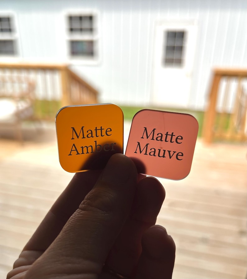 May include: Two square acrylic swatches with rounded corners. The left swatch is translucent amber with the text Matte Amber printed in black. The right swatch is translucent mauve with the text Matte Mauve printed in black. Both items are held up.