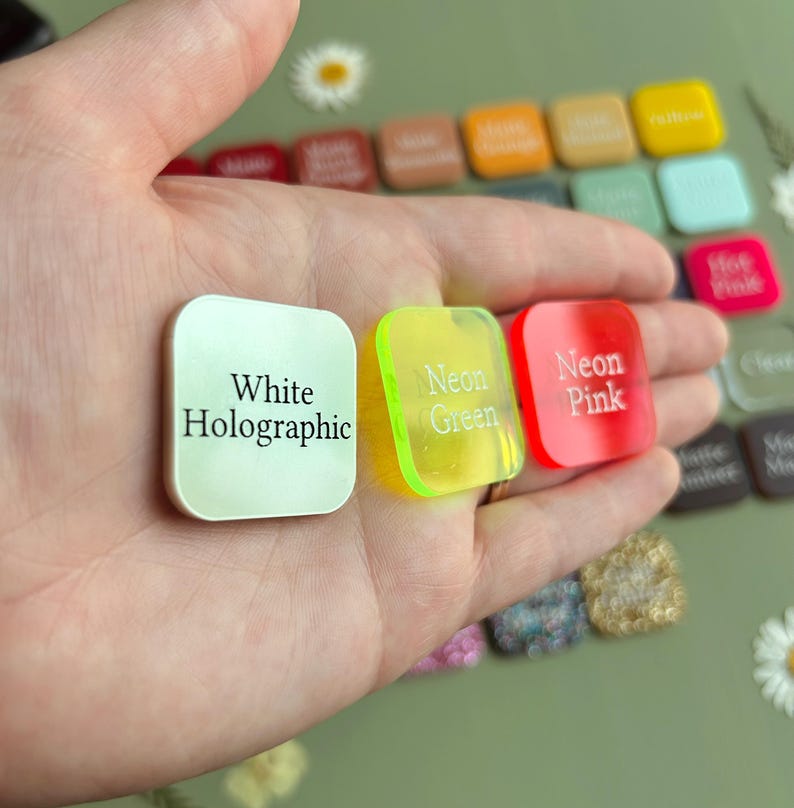 May include: A collection of small, square-shaped acrylic color swatches. Three pieces are held in the palm, labeled White Holographic, Neon Green, and Neon Pink. Additional swatches in various colors and finishes are arranged in the background.