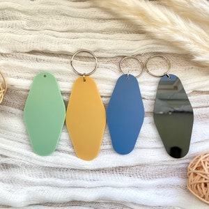 May include: Four elongated diamond-shaped keychains in matte light green, matte mustard yellow, matte blue, and a reflective black finish. Three of the keychains feature attached silver-toned metal split rings for keys, while the green one has a small hole at the top.
