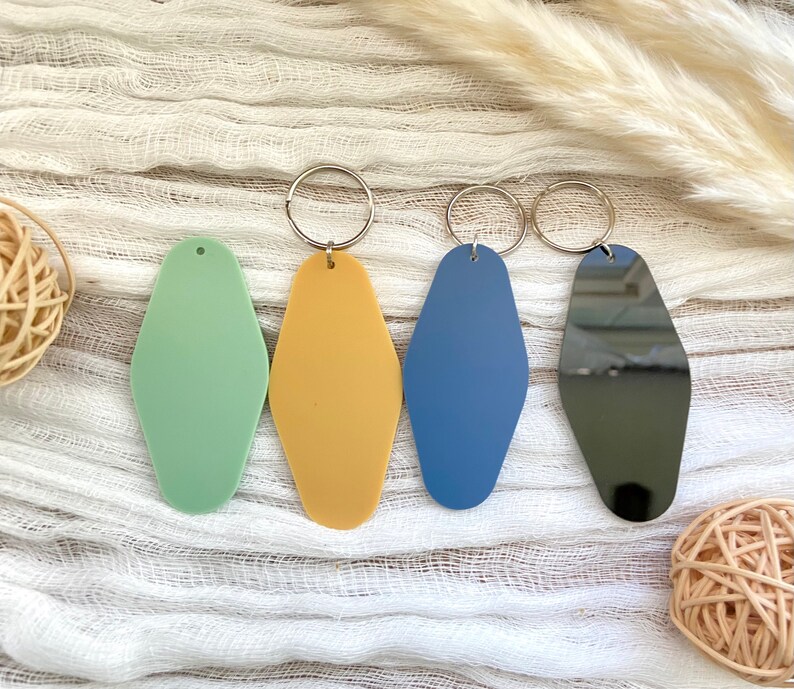 May include: Four elongated diamond-shaped keychains in matte light green, matte mustard yellow, matte blue, and a reflective black finish. Three of the keychains feature attached silver-toned metal split rings for keys, while the green one has a small hole at the top.
